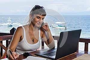 Young pretty woman freelancer writer working with laptop notepad and phone in front of blue tropical sea