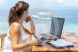 Young pretty woman freelancer writer working with laptop notepad and phone infront of blue tropical sea