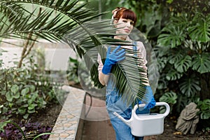Young pretty woman gardener, wearing jens overalls and blue rubber gloves, posing with water can, holding big palm leaf