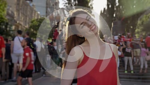 Young pretty woman with ginger hair standing on street during festival, looking in camera and smiling, crowd of fans