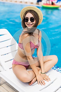 Young pretty woman with hat and sunglasses itting and relaxing in chaise lounge at the poolside