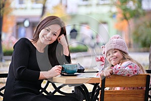 Young pretty woman having good time with her child daughter sitting at street cafe with hot drinks on sunny autumn day