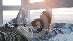 Young pretty woman in headphones relaxing with her lovely Maine Coon cat listens to music laying in bed by the window