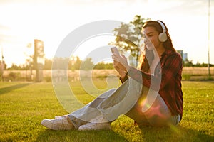 Young pretty woman in headphones uses a mobile phone for listening to music sitting on the grass