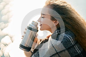 Young pretty woman holds a thermos and drinks tea wrapped in blanket over blur river background.