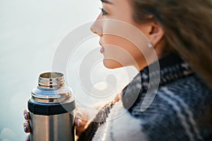 Young pretty woman holds a thermos and drinks tea wrapped in blanket over blur river background.