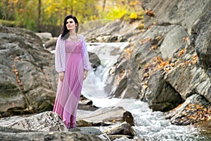 Young pretty woman in long fashionable dress standing near small mountain river with fast moving water