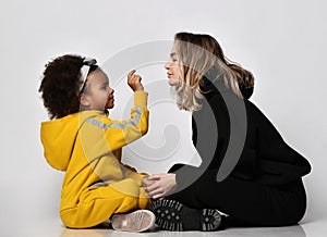 Young pretty woman mother in black clothing and curly cute mulatto daughter girl in sport jumpsuit sitting and communicating