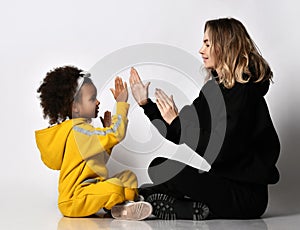 Young pretty woman mother and curly cute mulatto daughter girl in sport jumpsuit and sneakers sitting and playing hands