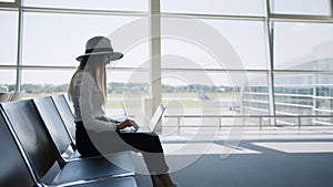 Young pretty woman printing on laptop sitting at airport. Girl tourist freelancer works and waits for flight in waiting