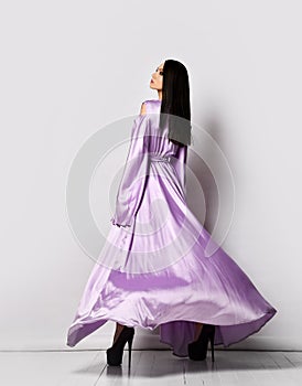 Young pretty woman in purple silky flying maxi dress and shoes standing backwards feeling light and soft after laser
