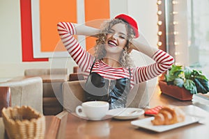 Young pretty woman in red beret having a french breakfast with coffee and croissant sitting at the cafe
