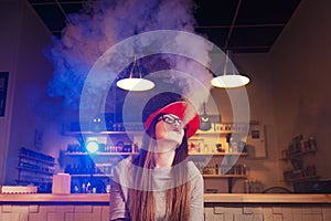 Young pretty woman in red cap smoke an electronic cigarette at the vape shop