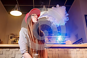 Young pretty woman in red cap smoke an electronic cigarette at the vape shop