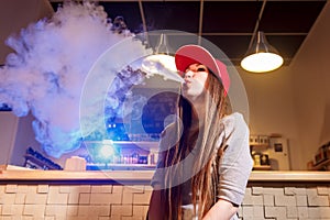 Young pretty woman in red cap smoke an electronic cigarette at the vape shop