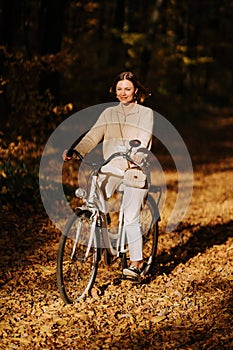 Young pretty woman riding vintage white bicycle in autumn park. Lady having fun on orange nature fall background.