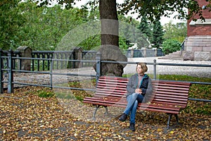 A young, pretty woman sits in a park on a bench, cross-legged, holding a telephone in her hand. Around are lying on the ground