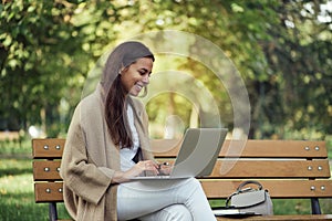 Young pretty woman sitting on bench at the park during summer day and using laptop.