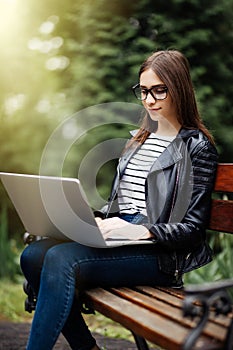 Young pretty woman sitting on bench in the park, using laptop computer, networking online on wireless. Student work on laptop in p