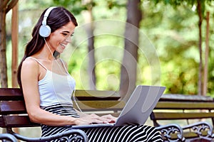 Young pretty woman with headphones sitting on bench in the park, using laptop computer