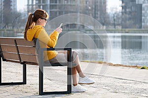 Young pretty woman sitting on a park bench browsing her smartphone outdoors on warm autumn day