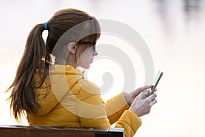 Young pretty woman sitting on a park bench browsing her smartphone outdoors on warm autumn day