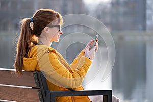 Young pretty woman sitting on a park bench browsing her smartphone outdoors on warm autumn day