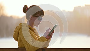 Young pretty woman sitting on a park bench browsing her smartphone outdoors in warm autumn evening.