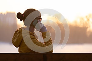 Young pretty woman sitting on a park bench talking on her smartphone outdoors in warm autumn evening
