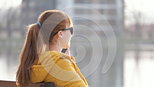 Young pretty woman sitting on a park bench talking on her smartphone outdoors in warm autumn evening.