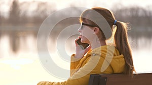 Young pretty woman sitting on a park bench talking on her smartphone outdoors in warm autumn evening.