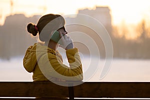 Young pretty woman sitting on a park bench talking on her smartphone outdoors in warm autumn evening