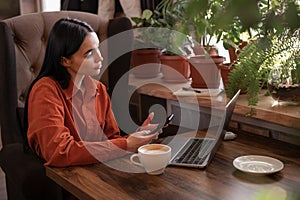 Young pretty woman sitting at table holding smartphone and using laptop computer, looking at mobile phone while remote
