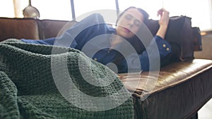 Young pretty woman sleeping on a brown leather sofa, she covered with a green plaid. The camera shoots from left to