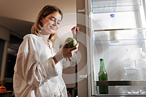 Young pretty woman standing on kitchen at night near opened fridge happily looking on green apple