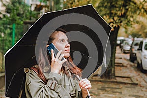 young pretty woman talking on the phone at city street hiding from rain under umbrella