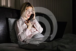 Young pretty woman talking phone while working in laptop at home in bed