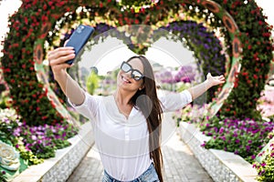 Young pretty woman tourist take selfie on the phone in miracle garden with colorfull frowers hearts