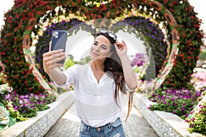 Young pretty woman tourist take selfie on the phone in miracle garden with colorfull frowers hearts