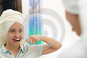 A young pretty woman with a towel on her head in a bright bathroom with a toothbrush in her hand brushes her teeth. Reflection in