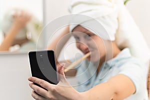 A young pretty woman with a towel on her head in a bright bathroom with a toothbrush and a smartphone in her hand brushes her teet