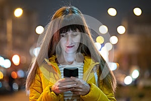 Young pretty woman using her mobile phone standing on city street at night outdoors