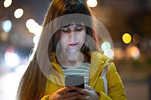 Young pretty woman using her mobile phone standing on city street at night outdoors