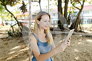 Young pretty woman using tablet and riding swing on sand, wearing jeans sundress.