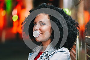 Young pretty woman with very curly afro hair inflates a bubble ball of white chewing gum at night. Trendy girl having