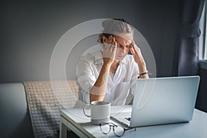 Young pretty woman in a white shirt sits at a table in front of a laptop, her eyes and head ache, workaholism, stress burnout