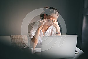 Young pretty woman in a white shirt sits at a table in front of a laptop, her eyes and head ache, workaholism, stress burnout photo