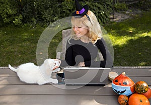 Young pretty woman in witch hat. Sits at table in garden, works at laptop