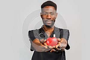 Young professional african doctor cardiologist with stethoscope holding red heart in hand, standing against gray background.