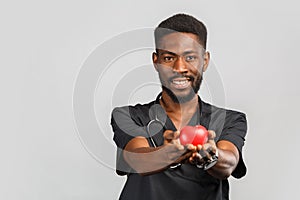 Young professional african doctor cardiologist with stethoscope holding red heart in hand, standing against gray background.